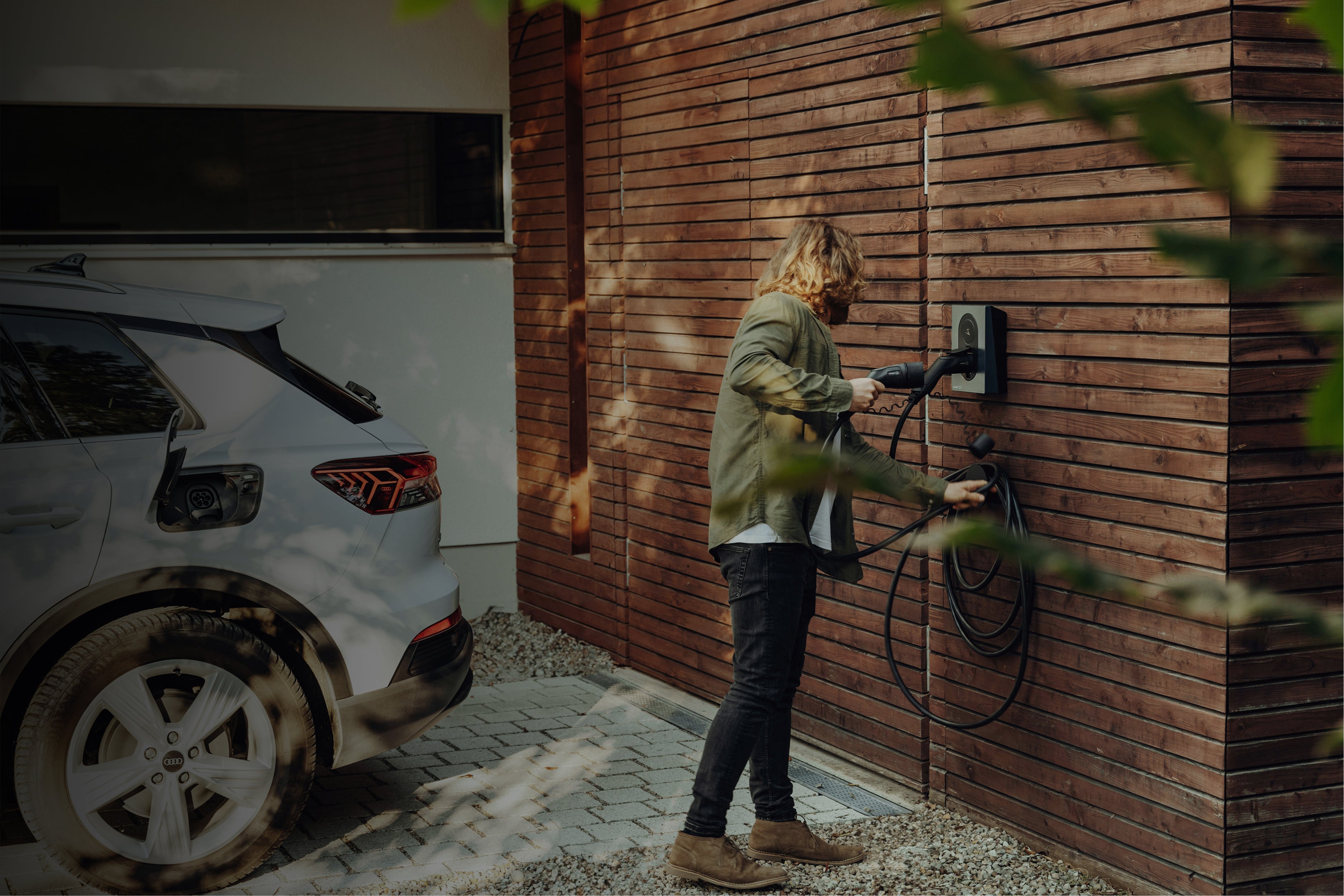 Person charging a white electric car at a charging station on a wooden wall.